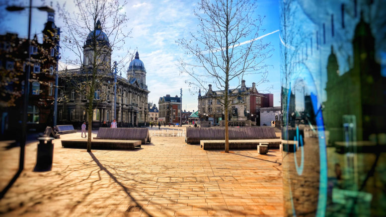 City centre buildings with a blue sky and light coloured paving, with trees and outdoor seating