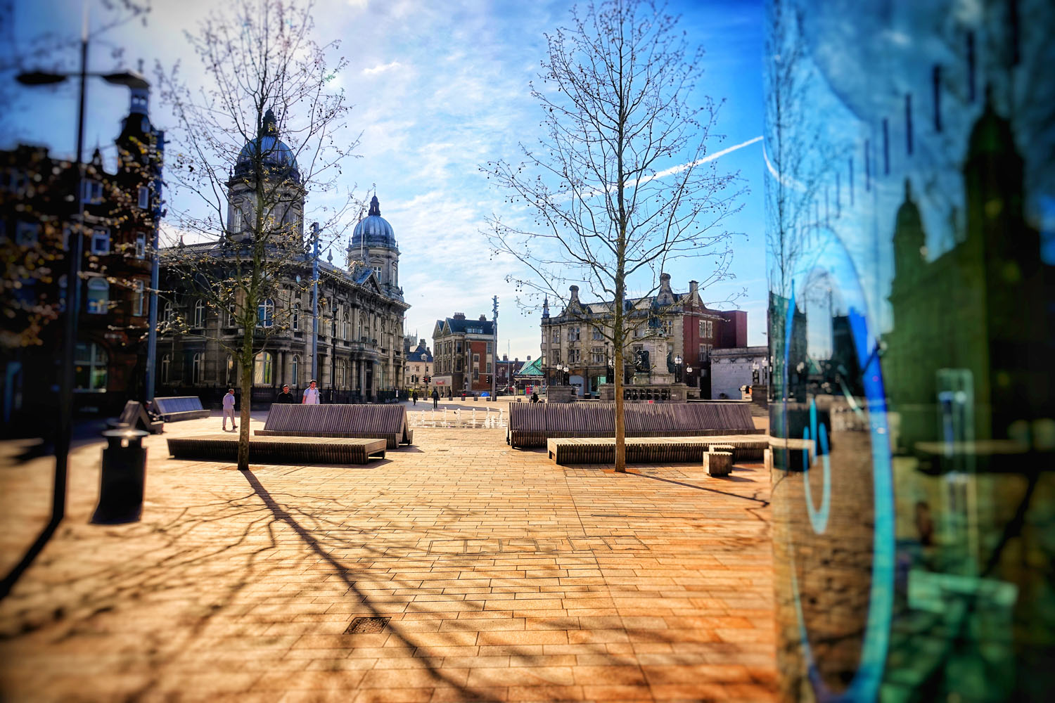City centre buildings with a blue sky and light coloured paving, with trees and outdoor seating