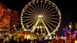 A shot of a fairground at night. Different coloured neon-lit stalls are in the foreground. A big wheel, lit white, takes up most of the picture