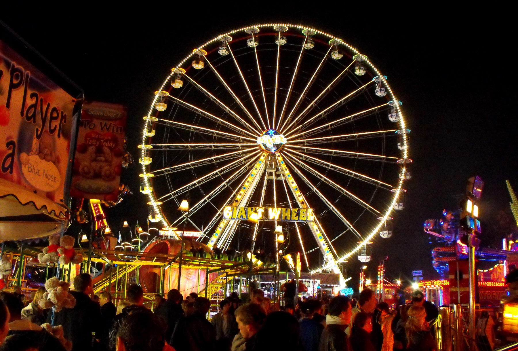 A shot of a fairground at night. Different coloured neon-lit stalls are in the foreground. A big wheel, lit white, takes up most of the picture