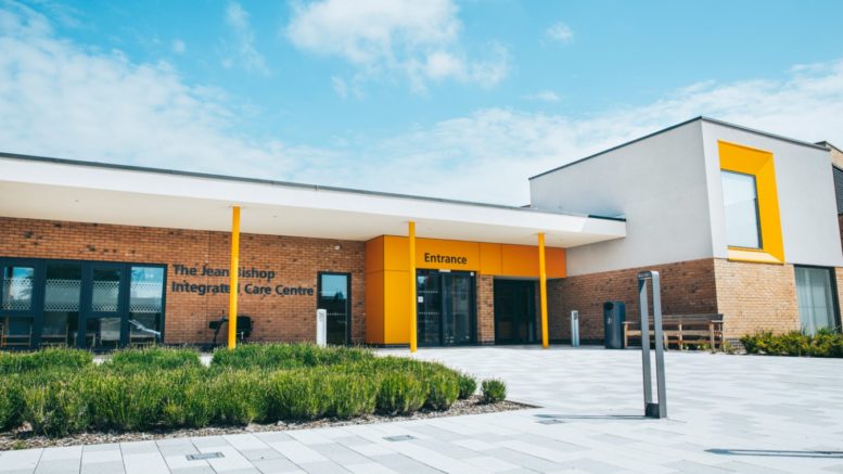 The Jean Bishop Integrated Care Centre. The name is on the side of the building. It has a yellow frontage, with green shrubbery and the pavement around the building is grey.