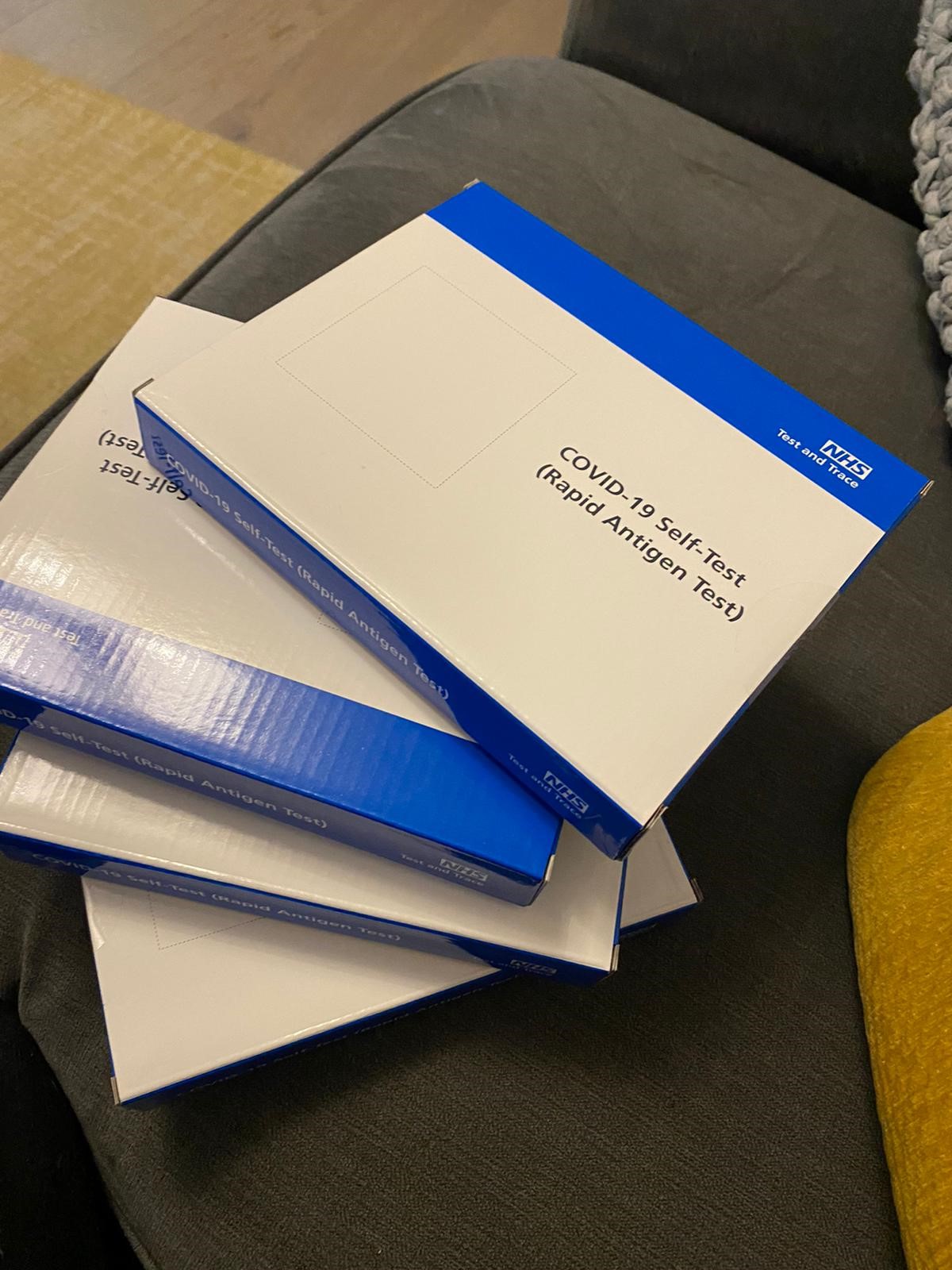 boxes containing lateral flow tests are stacked on a table. The boxes are white and blue and have an NHS logo