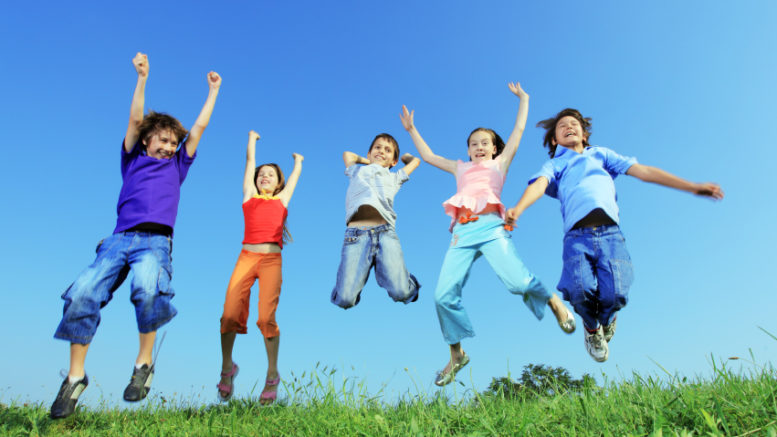 five children jump joyfully outside, in sunny weather. The picture is taken from ground-level, looking up at them