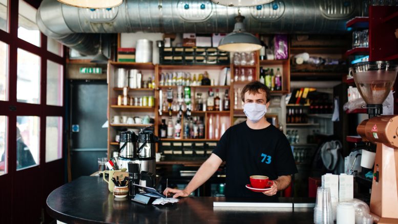 A person serves coffee indoors. They wear a facemask