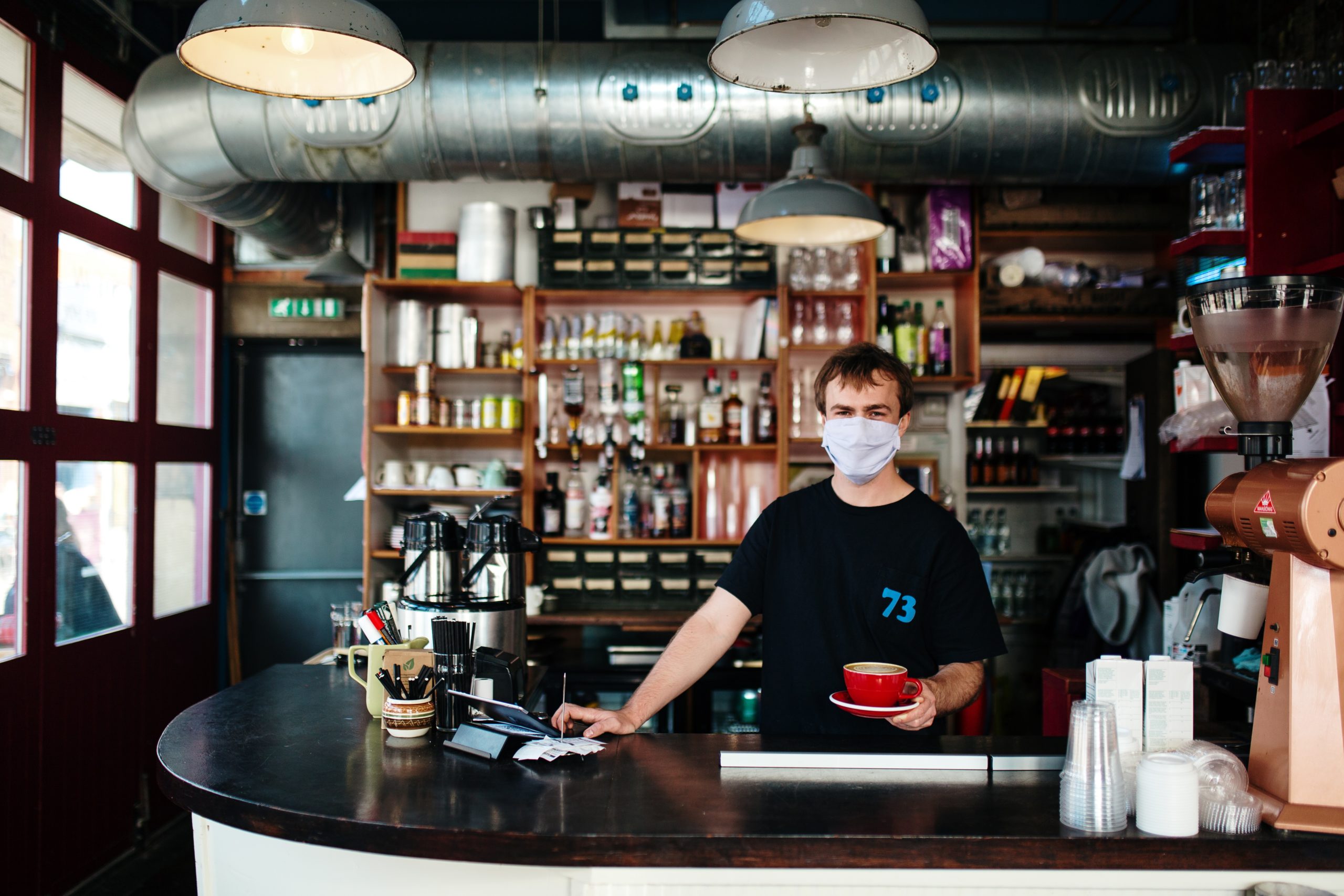A person serves coffee indoors. They wear a facemask