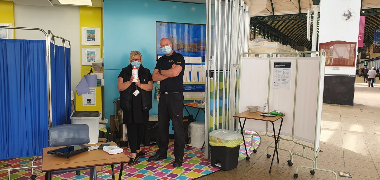 two people wearing masks stand in a pop-up Coronavirus testing booth. It is inside a rail interchange