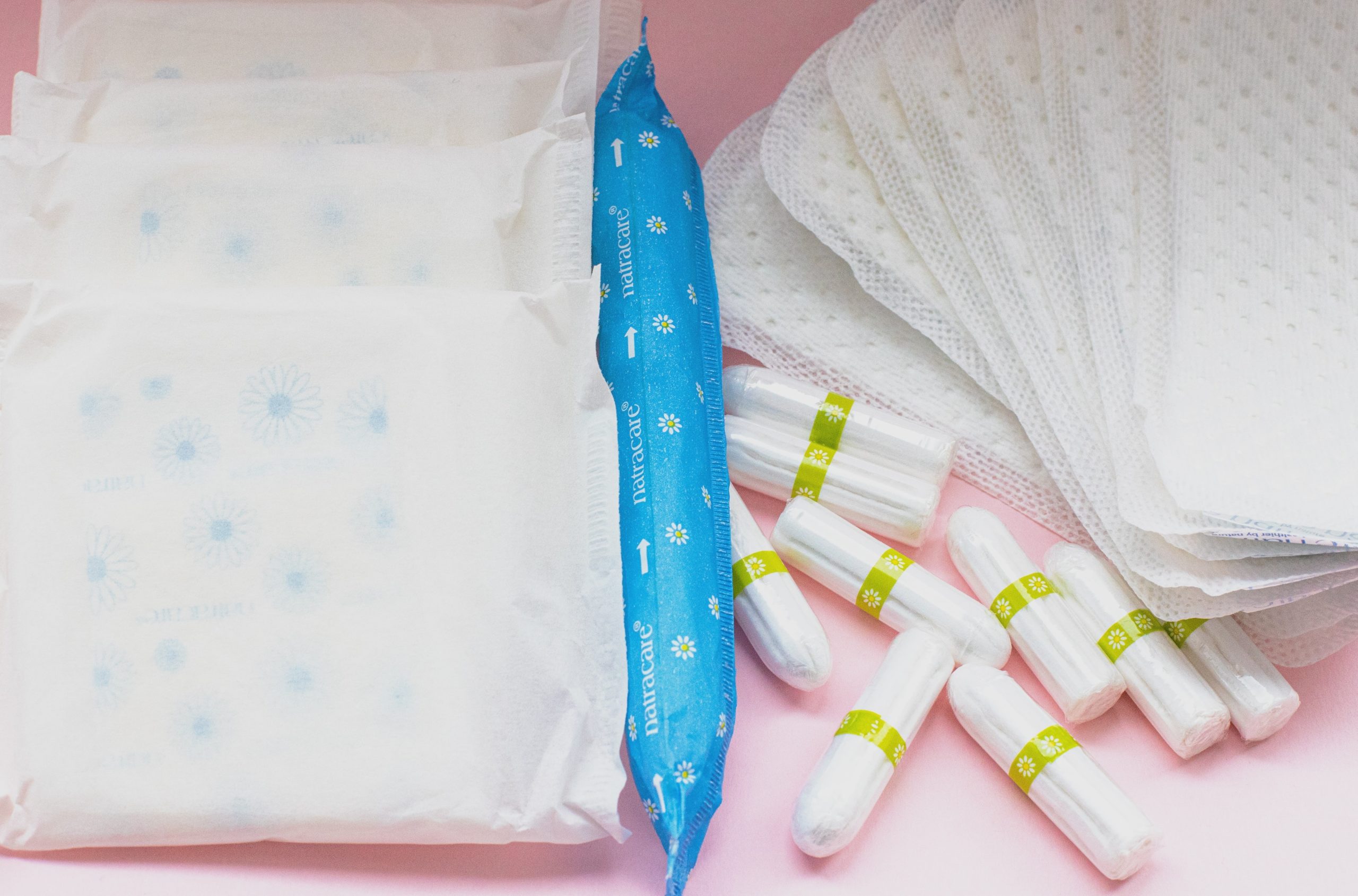 a selection of single-use period products, including pads and tampons. They are in their packaging, on a pick background.