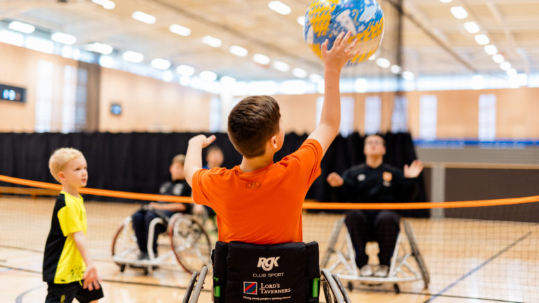 a group of young people in wheelchairs play volleyball in a brightly-lit sportshall