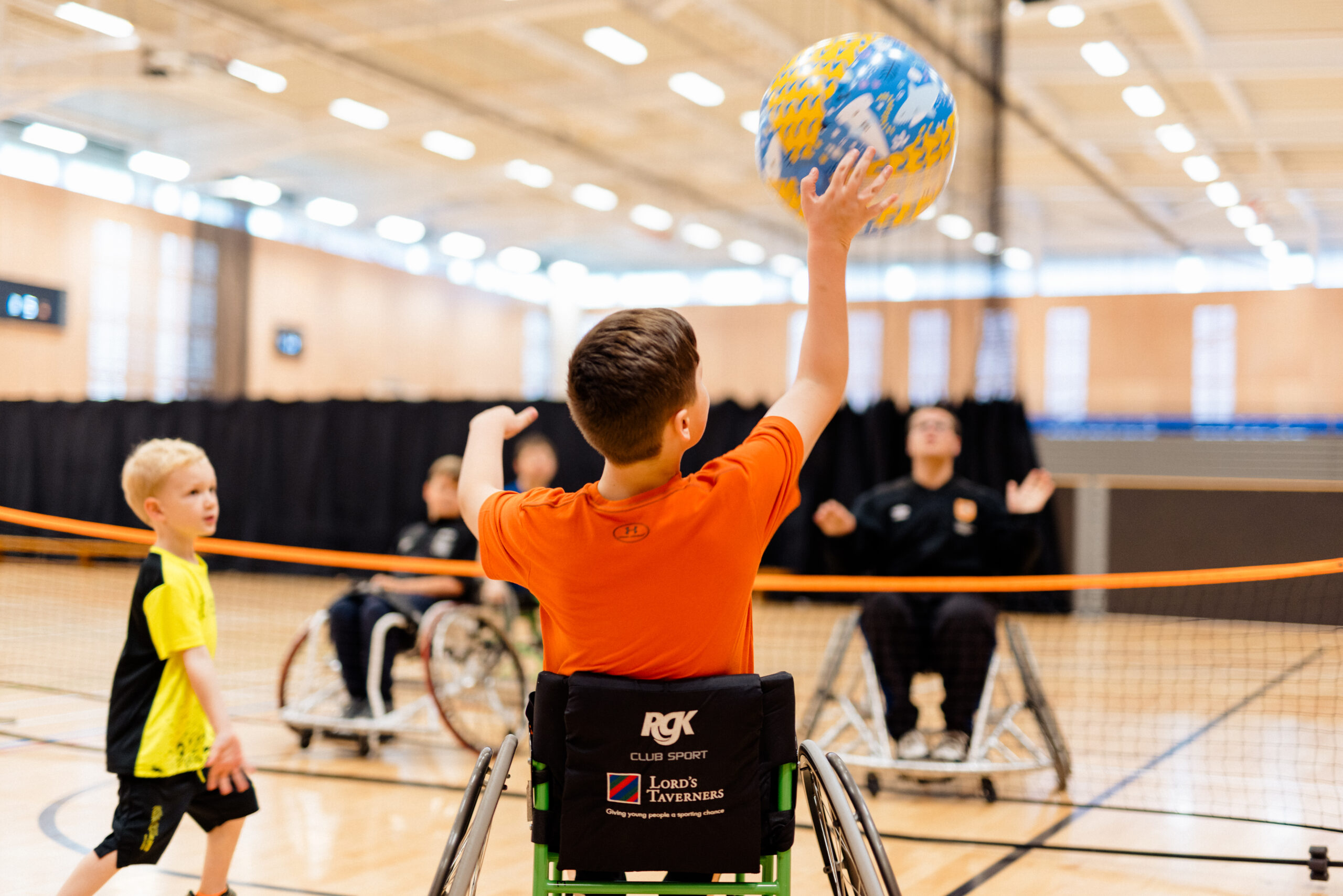 a group of young people in wheelchairs play volleyball in a brightly-lit sportshall
