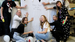 A group of students stand together, next to a display about White Ribbon Day. They are preparing to make palm prints on a plain white sheet.