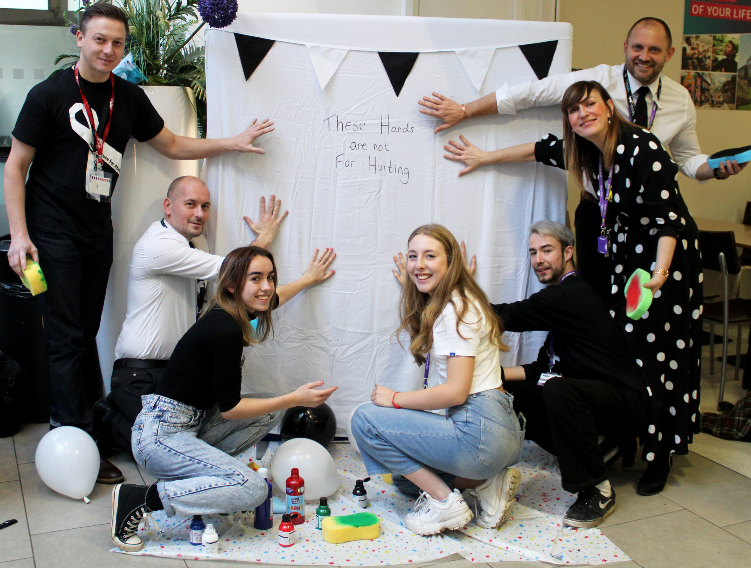 A group of students stand together, next to a display about White Ribbon Day. They are preparing to make palm prints on a plain white sheet.