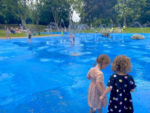 A large water play area with fountains and jets and a blue painted surface. Children playing in background with 2 children in foreground.