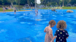 A large water play area with fountains and jets and a blue painted surface. Children playing in background with 2 children in foreground.