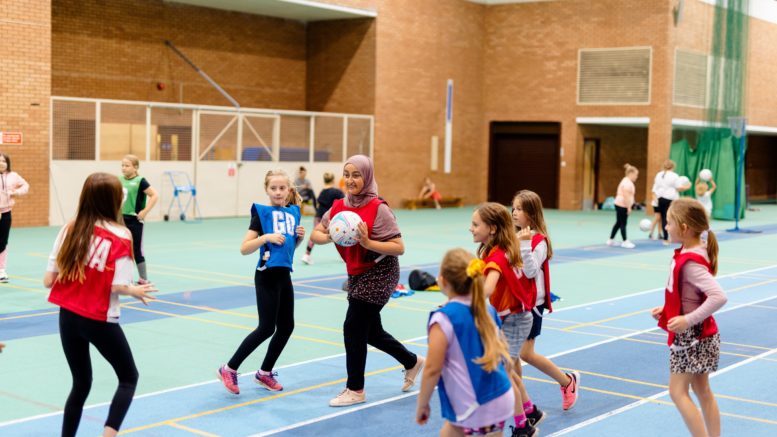 a group of young girls are playing netball in a sportshall. A girl wearing a hijab is preparing to make a pass