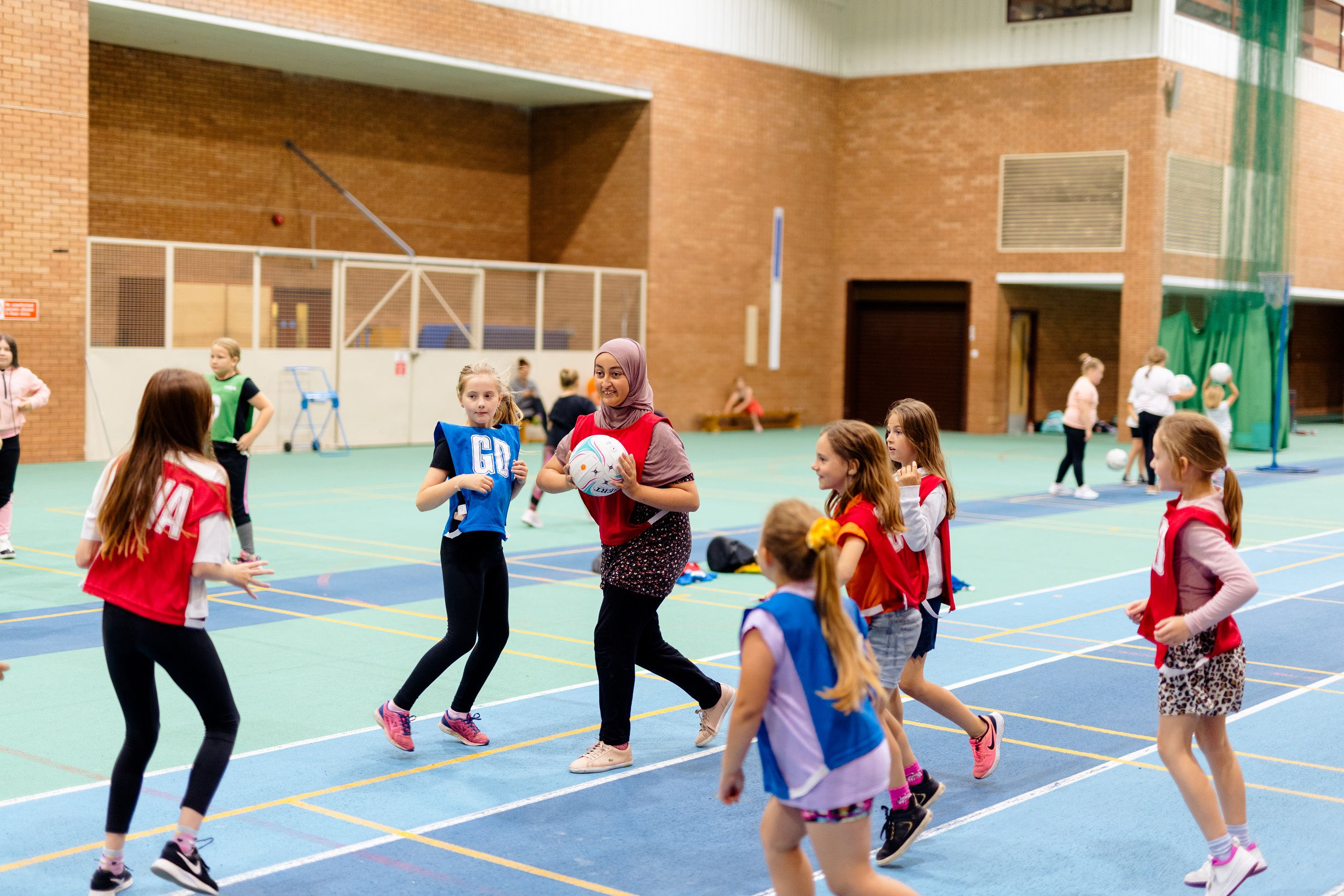 a group of young girls are playing netball in a sportshall. A girl wearing a hijab is preparing to make a pass