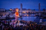 Mo and The Red Ribbon outdoor performance. A giant puppet walks through an outdoor crowd at night. A bridge across an inner city river full of boats can be seen in the background.