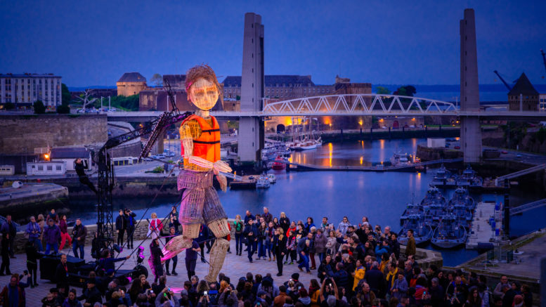 Mo and The Red Ribbon outdoor performance. A giant puppet walks through an outdoor crowd at night. A bridge across an inner city river full of boats can be seen in the background.
