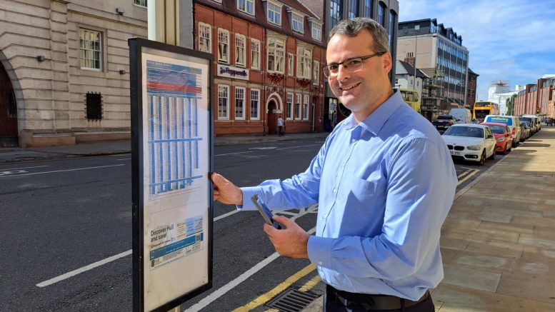 ouncillor Mark Ieronimo standing at a QR-enabled bus stop on Alfred Gelder Street in Hull, holding a smartphone in his hand