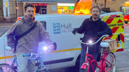 Two male cyclists on bikes in front of a Hull CIty Council van by the side of a road at dusk. The bikes have lights on their handlebars.