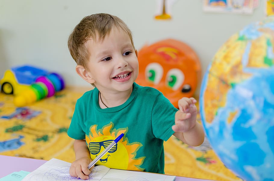 A boy smiling and playing with nursery toys, he wears a green t shirt. He sits at a table touching a globe.
