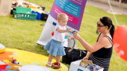 a woman and a toddler sit on blankets on the ground at a summer community event. The woman is showing the toddler handheld instruments with bells on.