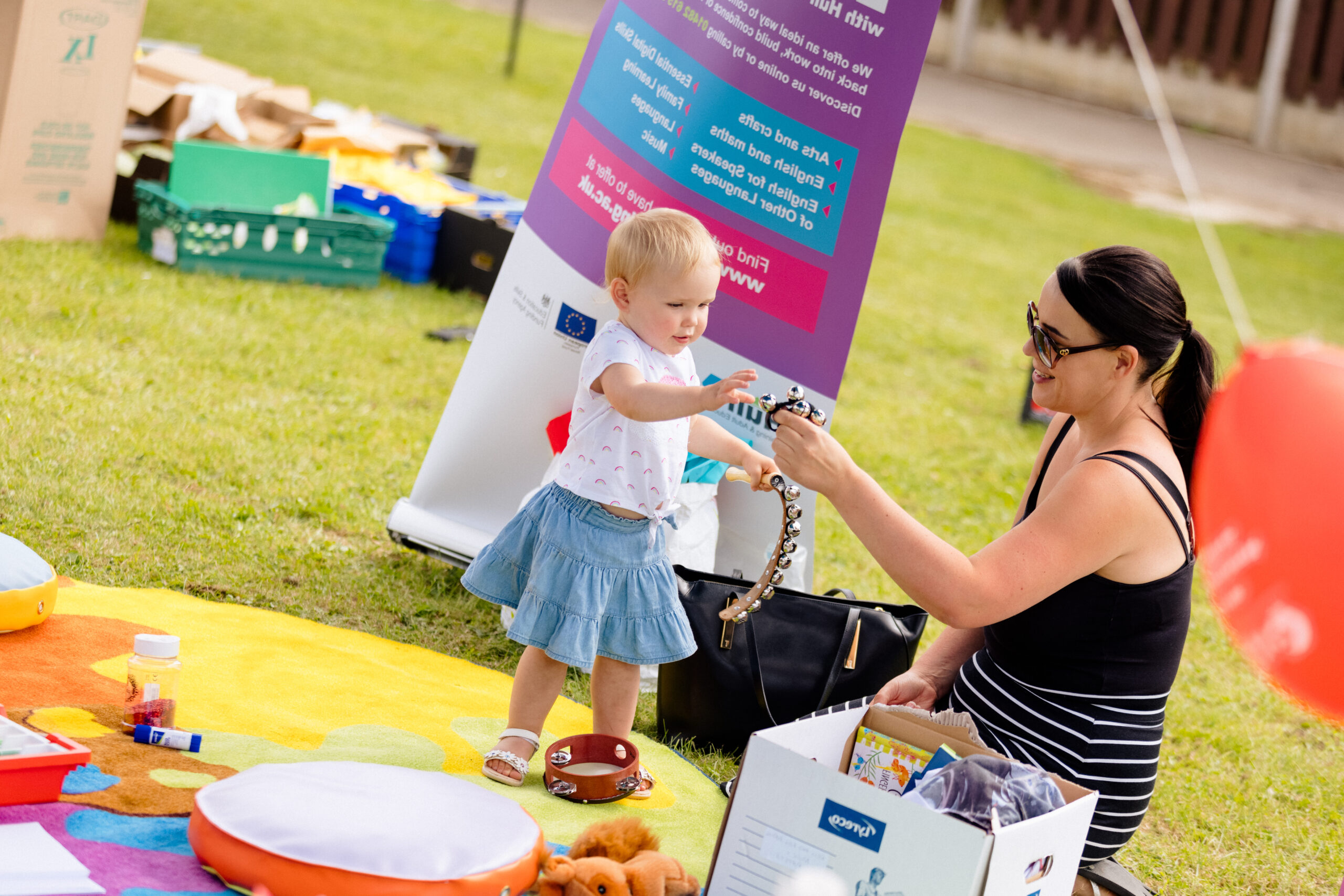 a woman and a toddler sit on blankets on the ground at a summer community event. The woman is showing the toddler handheld instruments with bells on.