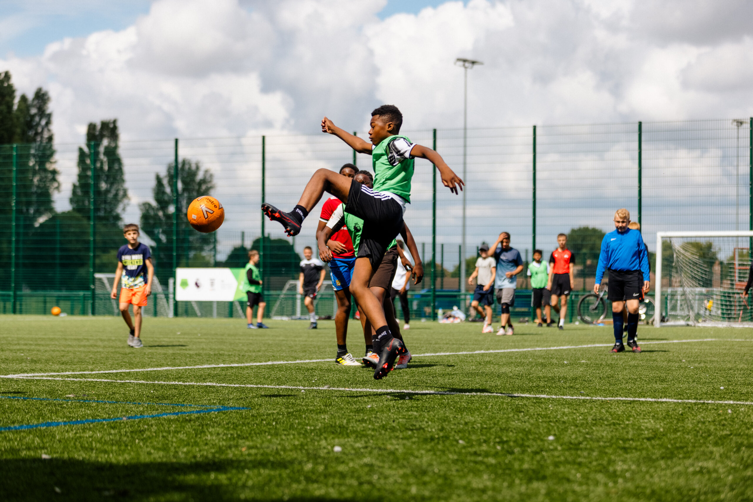 A group of children are playing football outside in the sun. a boy in the centre of the photo is lifting his leg high in the air to kick a ball