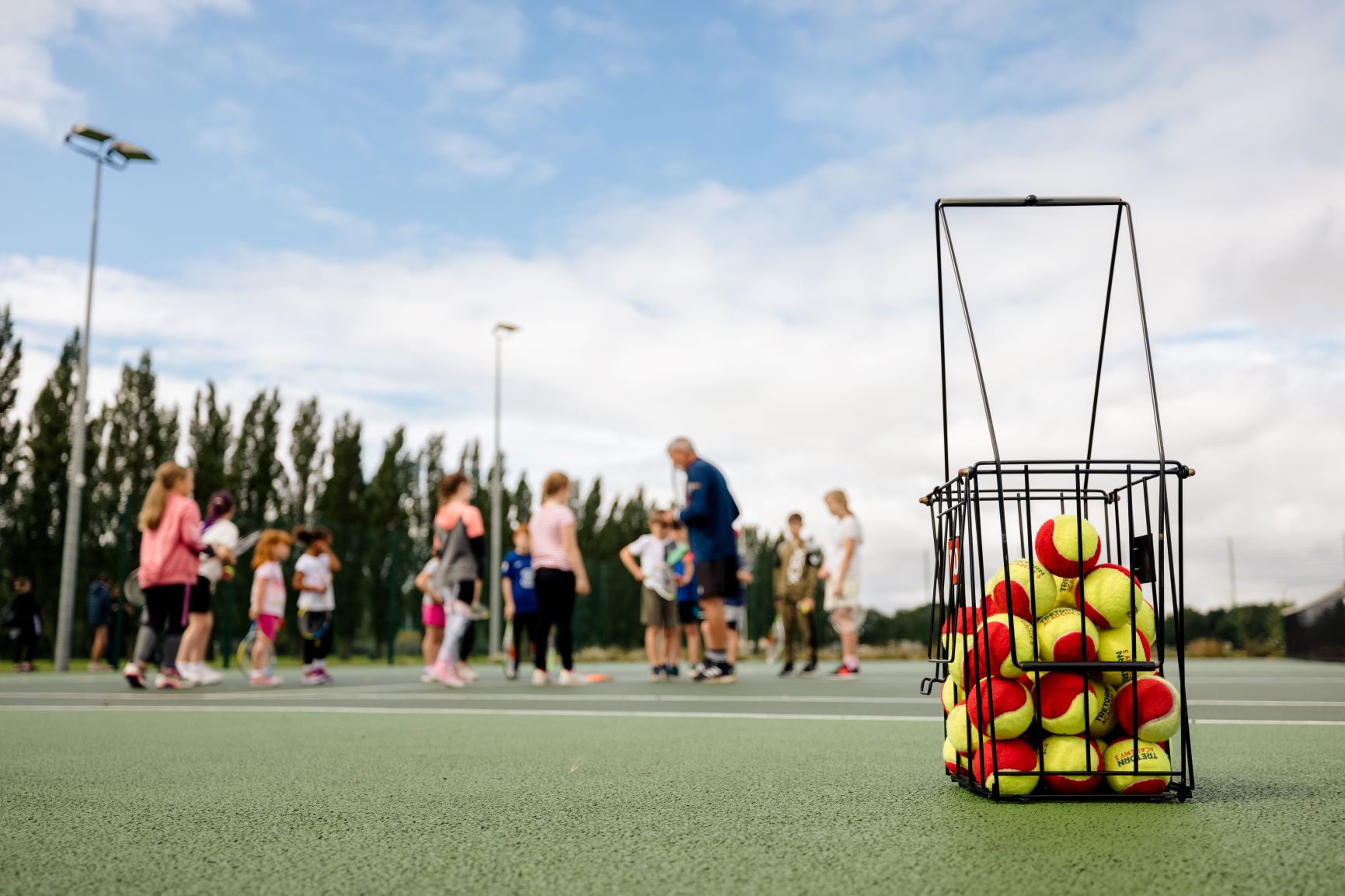 A basket of tennis balls with a group of young tennis players in the background