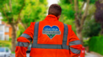 A Hull City Council worker seen from behind wearing a high-visibility jacket emblazoned with the "Love Your Neighbourhood" logo. In the background is a spring scene in a British street