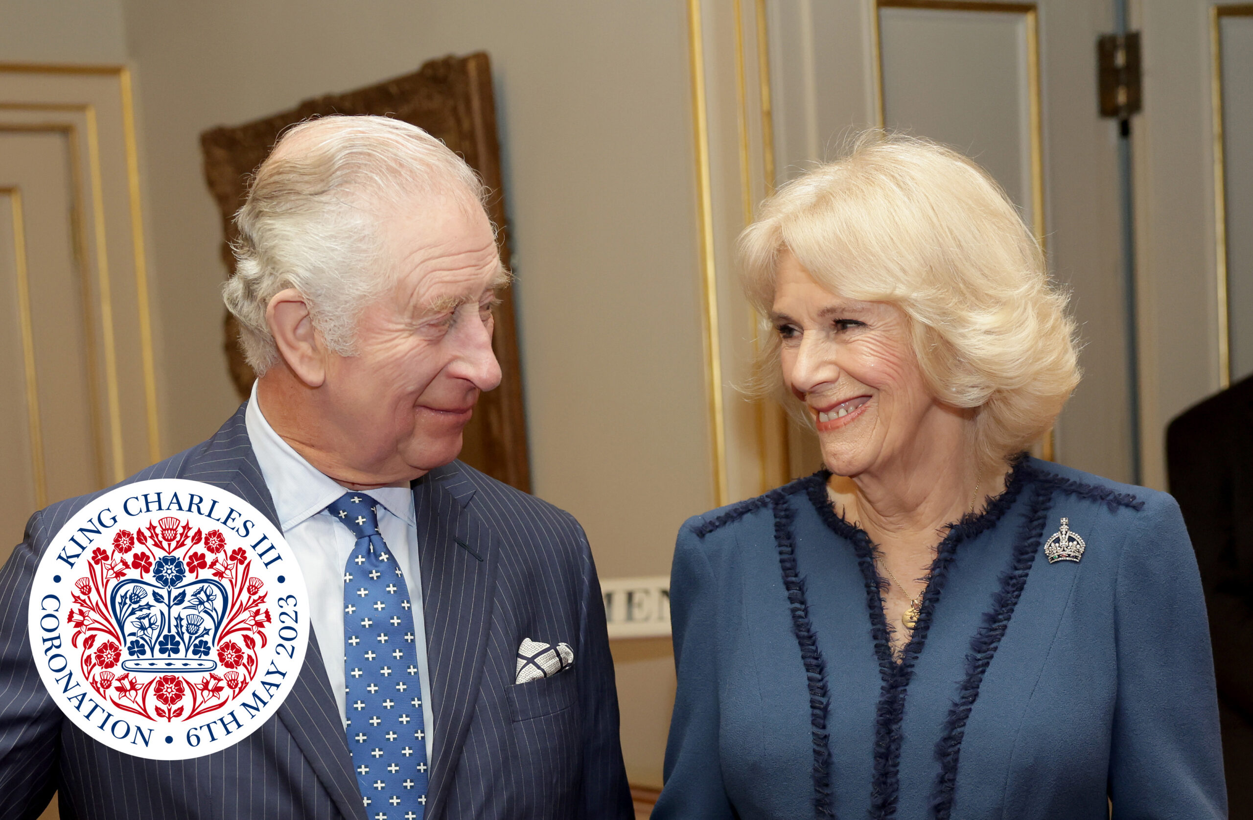 King Charles III and Queen Consort Camilla smiling with royal coronation logo in bottom left corner