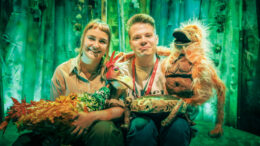 Two performers from the Whispering Jungle sit together on the set, which looks like a jungle, complete with green lighting. They both hold puppets which are used in the performance