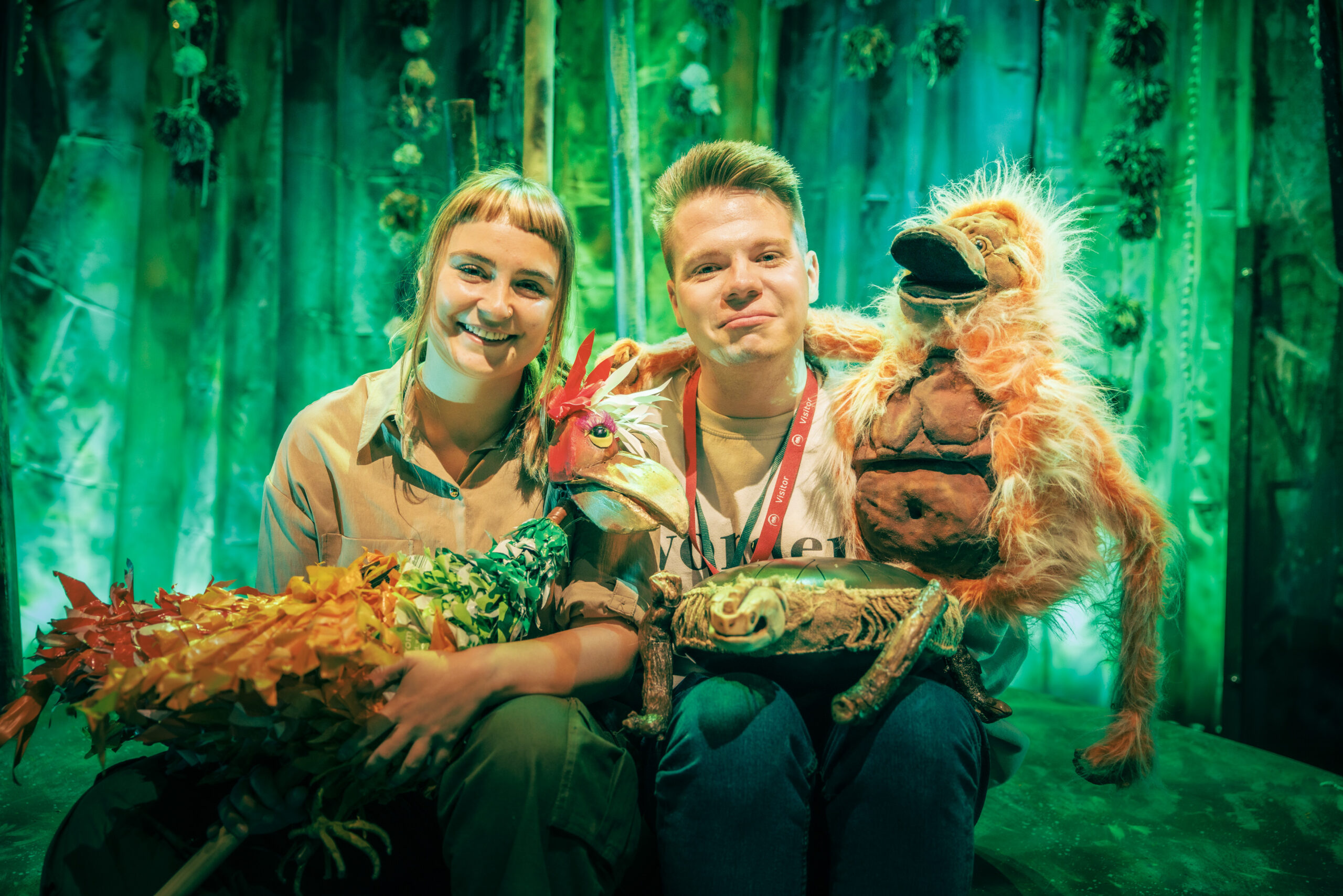 Two performers from the Whispering Jungle sit together on the set, which looks like a jungle, complete with green lighting. They both hold puppets which are used in the performance