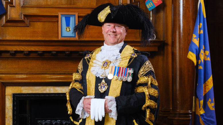 The Lord Mayor stands in his regalia in front of a dark wooden fireplace