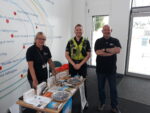 A photo taken at an event for Not Where I Live Week in 2022. Three people stand inside a mobile surgery. There is a table of crime prevention merchandise with them. One person is a police officer in uniform.