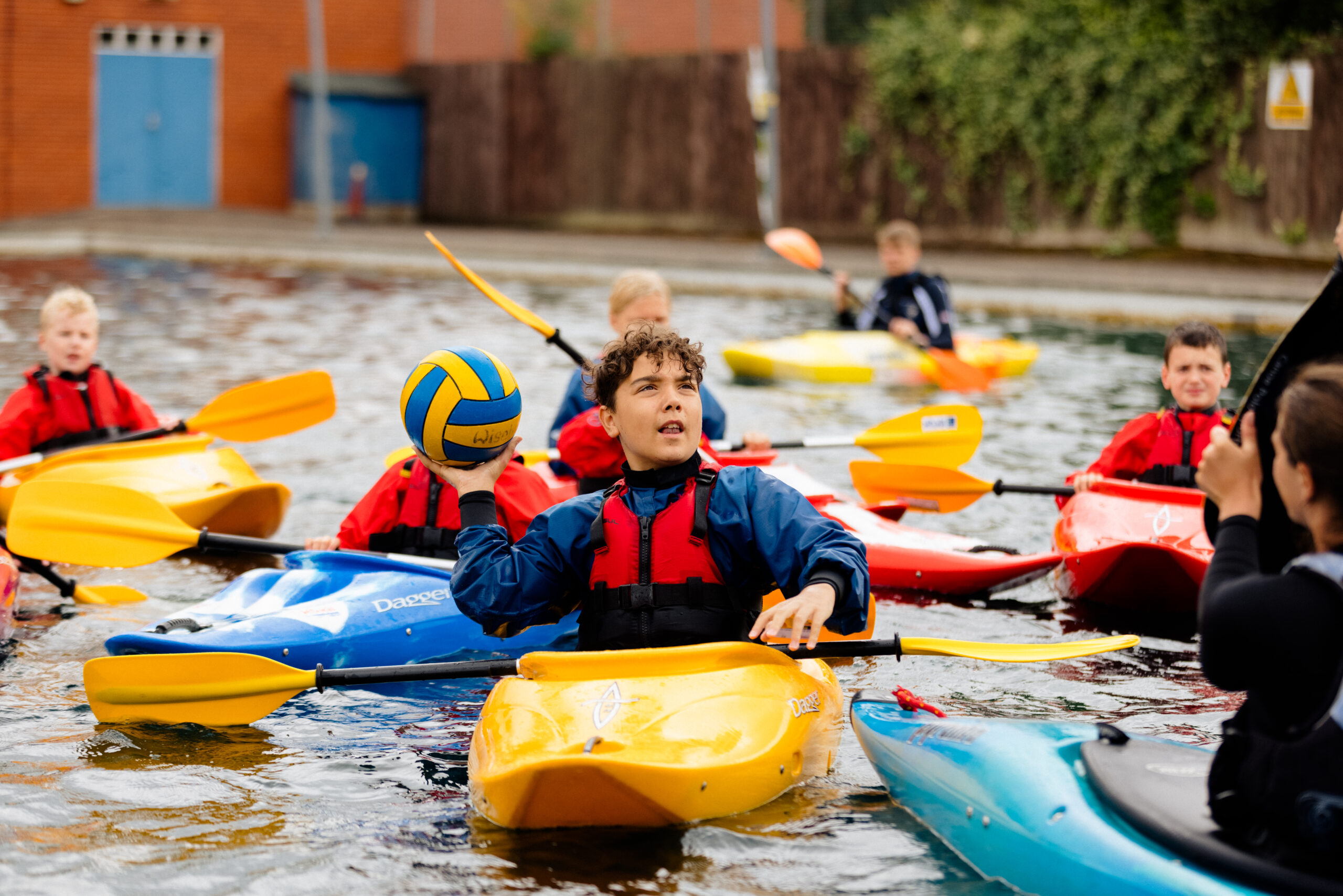 young people are playing a ball game, while using kayaks in an outdoor pool. The kayaks are bright yellow and blue