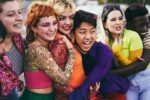 a group of young people wearing brightly-coloured clothes stand close together in a group. They are smiling and hugging.