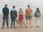 A family stood on a beach facing the sea, with their pet dog. Their foster child, a girl, is stood in the middle.