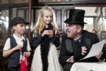 A man talks to 2 children, all in Victoria dress