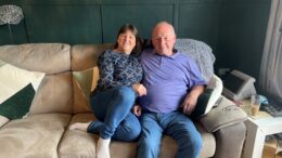 Janet and Clive are sat on a sofa together smiling.