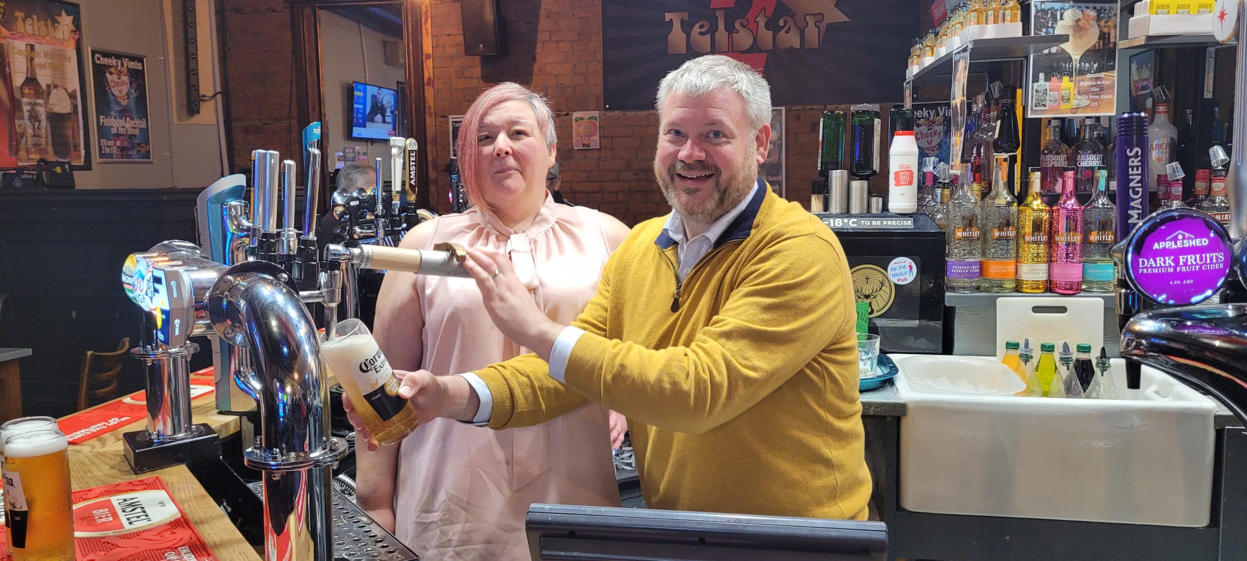 two people stand behind a bar. One pulls a pint. They are both smiling.