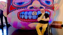 Artist Jason sits in a wheelchair in front of a painted colourful mask