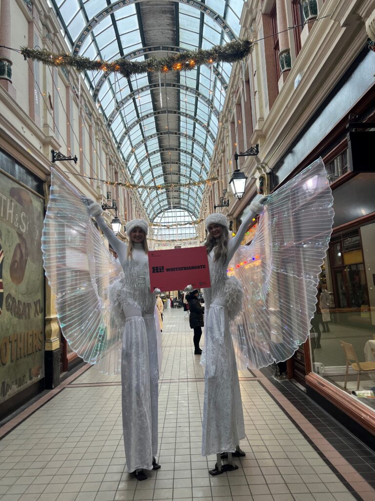 2 women dressed in white as angels on Stilts holding a HI! Whitefriargate sign