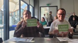 Two men sat smiling with signs saying 'I want to speak'.