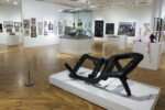 A landscape image of an gallery with frames on the wall and a black abstract sculpture in the foreground.