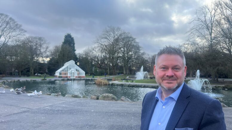 an image of Cllr Dave McCobb at Pearson Park. He is in the foreground and is smiling. Behind him you can see the park's duck pond, fountains and conservatory.