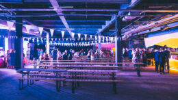 An image of an industrial style event space with benches, tables and bunting. With people mingling in the background.