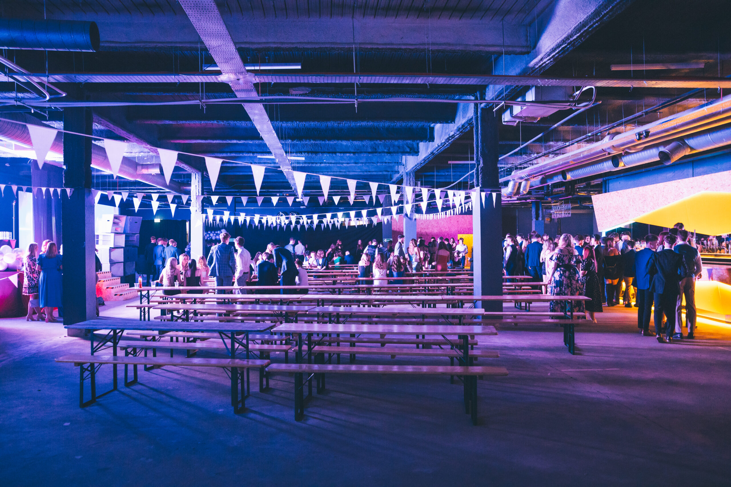 An image of an industrial style event space with benches, tables and bunting. With people mingling in the background.