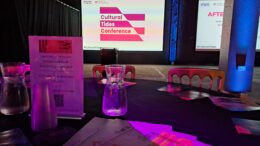 A picture of a table in the foreground with chairs facing a stage with a large screen with pink and white Cultural Tides logo