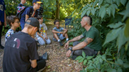 an adult guide and a group of children sit in sunny woodland in the summer. The guide is speaking to the children