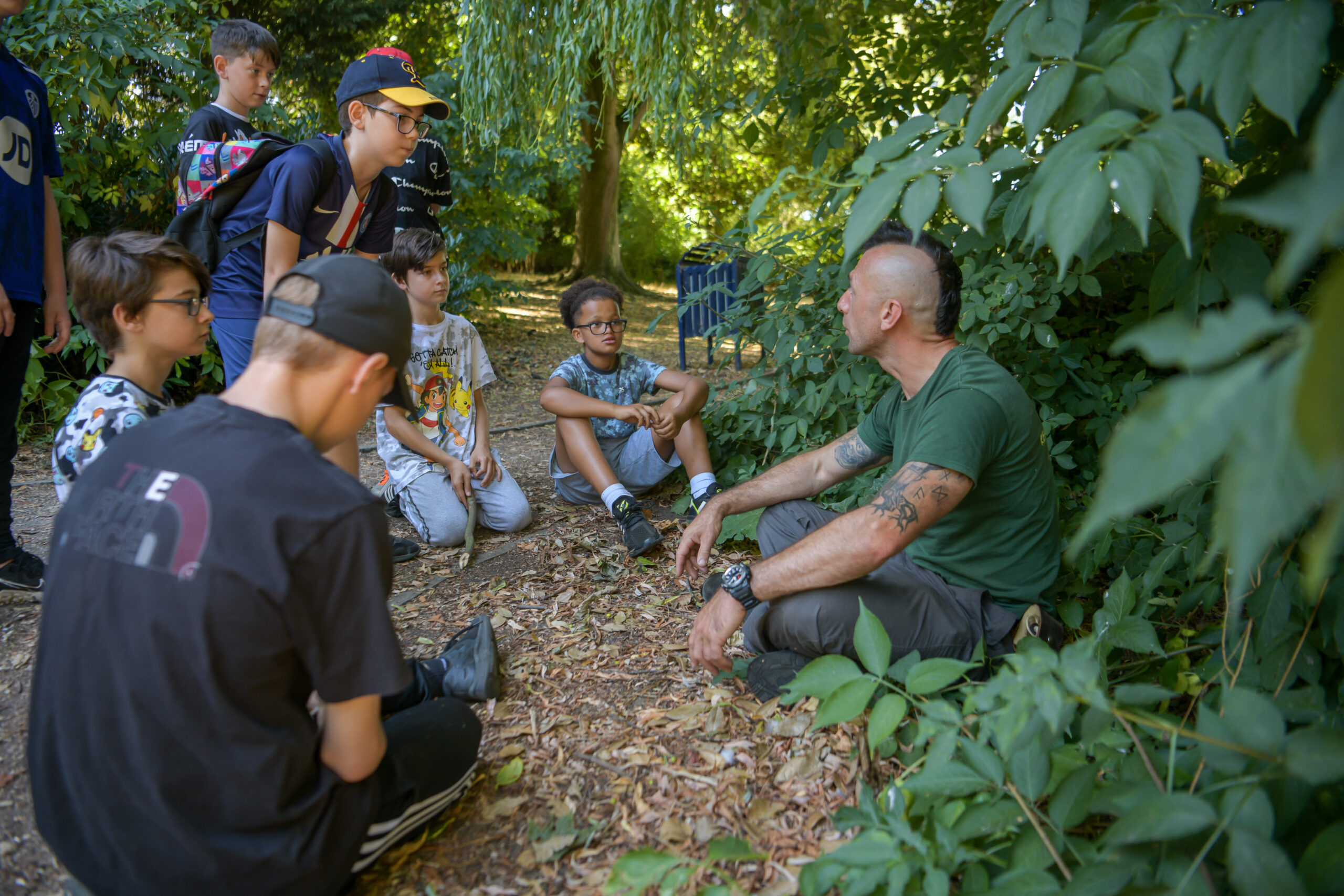 an adult guide and a group of children sit in sunny woodland in the summer. The guide is speaking to the children